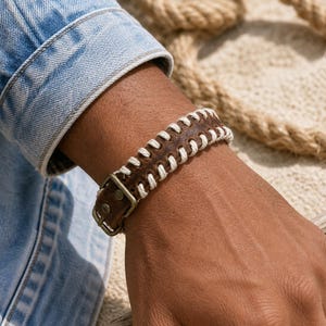 Pode incluir: Pulseira de couro castanho com costuras brancas e fivela de metal. A pulseira é usada no pulso, com uma manga de jaqueta jeans ao fundo. A pulseira tem aproximadamente 2,5 cm de largura.