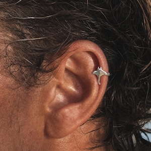 Peut inclure: Une boucle d'oreille en forme de raie manta, de couleur argentée, avec une finition métallique lisse et polie. Le bijou est placé sur le cartilage supérieur de l'oreille, les ailes de la raie manta s'étendant horizontalement sur l'oreille.
