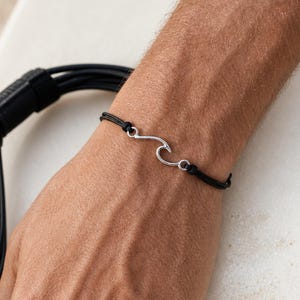 May include: A black cord bracelet with a silver wave design. The bracelet is on a person's wrist, with a surfboard and leash in the background. The wave design is a simple, elegant shape.