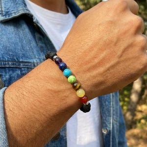 May include: A beaded bracelet featuring a variety of colorful gemstones and black lava beads. The bracelet has a mix of colors including red, orange, yellow, green, blue, and purple. The bracelet is worn on a wrist.