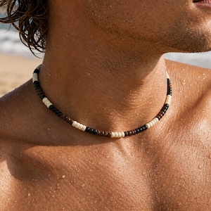 Peut inclure: Un collier de perles composé d'un motif répétitif de petites perles cylindriques marron foncé et crème. Le collier se porte près du cou et est constitué de perles uniformes, lisses et en forme de disque disposées en une séquence régulière.