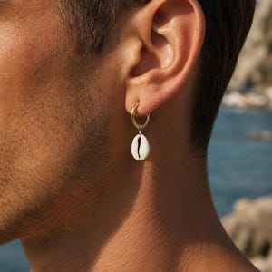 Peut inclure: Une boucle d'oreille créole dorée avec une petite breloque en coquillage cauri blanc suspendue. Le coquillage présente une forme ovale allongée naturelle avec une ouverture dentelée sombre courant verticalement au centre de la surface blanche.
