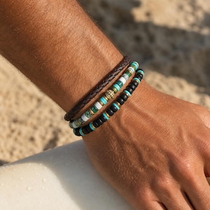Op de afbeelding: Drie armbanden op een pols. De bovenste armband is gevlochten bruin leer. De middelste armband heeft witte, turquoise en gouden kralen. De onderste armband heeft zwarte kralen met gouden accenten. Achtergrond is een zandstrand.