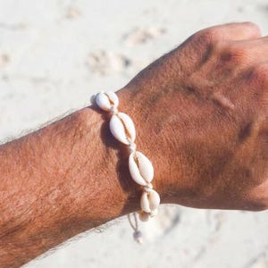 Puede incluir: Pulsera de conchas marinas naturales con cuatro conchas de cauri blancas unidas con un cordón beige. La pulsera se muestra en el brazo de una persona, con una playa de arena de fondo. Las conchas son ovaladas y tienen una textura suave y brillante.