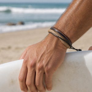 May include: A multi-strand bracelet featuring a dark brown leather band, a black leather cord, and a textured beige braided rope strand, all secured together with a black adjustable sliding knot closure on the side of the wrist.