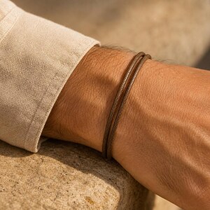 Könnte beinhalten: Ein zweisträngiges braunes Kordelarmband mit glatter, abgerundeter Textur. Das Armband wird am Handgelenk getragen und befindet sich direkt unter der Manschette eines langärmeligen, hellbeigen Leinenhemds, was ein schlichtes Design zeigt.