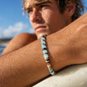 May include: A beaded bracelet featuring light blue, brown, and white beads. The bracelet is made of natural materials and has a casual, beachy aesthetic. The bracelet is shown on a person's arm.