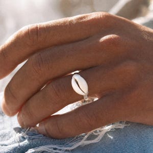 Könnte beinhalten: Ein Ring mit einem weißen Kaurimuschel-Mittelstück, das durch dünnen, silberfarbenen Draht befestigt ist. Die Muschel hat eine glatte, ovale Form mit einem zentralen Längsschlitz, und der Draht ist um das Band gewickelt, um die Muschel zu halten.