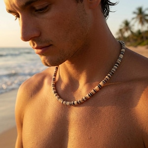 Puede incluir: Un collar de cuentas con segmentos marrones claros y oscuros alternados. El collar se lleva alrededor del cuello. El fondo muestra una escena de playa con palmeras y el océano. Un accesorio natural y terroso.