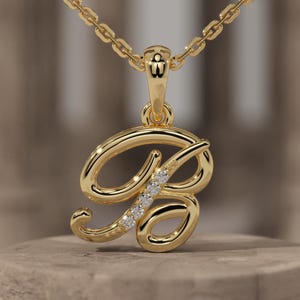 May include: A gold-colored pendant necklace featuring the letter "B" in a cursive script, embellished with small, sparkling stones. The pendant hangs from a delicate chain. The brand name "BREELIQ" is visible on the base.