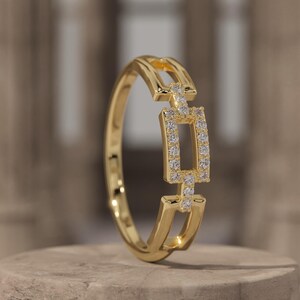 Puede incluir: Un anillo de oro con un elemento central rectangular con diamantes. El anillo tiene un diseño único con una serie de formas rectangulares conectadas. El anillo se muestra sobre una superficie de piedra con el nombre de la marca "BREELIQ".