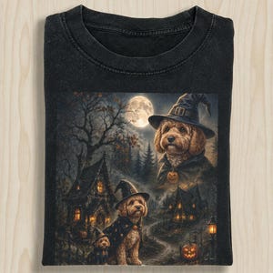 May include: A black t-shirt featuring a graphic print of three golden-colored dogs wearing witch hats and capes. The scene depicts a dark, spooky forest with a full moon, two rustic houses, and a glowing jack-o'-lantern in a nighttime setting.