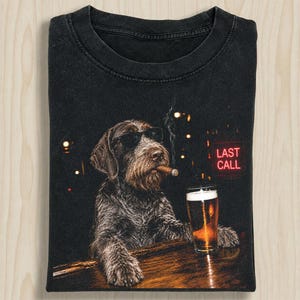 May include: A black t-shirt featuring a graphic of a wire-haired dog wearing sunglasses, smoking a cigar, and resting its paws on a wooden bar next to a glass of beer. A red neon sign in the background displays the text LAST CALL in capital letters.