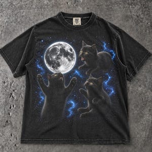 May include: A black short-sleeve t-shirt featuring a graphic print of three black kittens reaching toward a large, detailed full moon set against a dark blue starry night sky. The shirt has a crew neckline and a Comfort Colors brand label at the neck.