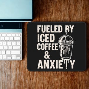 Puede incluir: Alfombrilla de ratón rectangular negra con el texto "Fueled by Iced Coffee & Anxiety" en blanco. Un gráfico de una taza de café helado está al lado del texto. Un teclado y un monitor de ordenador están en el fondo.