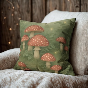 May include: Square decorative pillow featuring a whimsical illustration of red and white spotted mushrooms set against a green background with ferns. The pillow has a soft, inviting appearance, perfect for adding a touch of nature-inspired decor to any room.