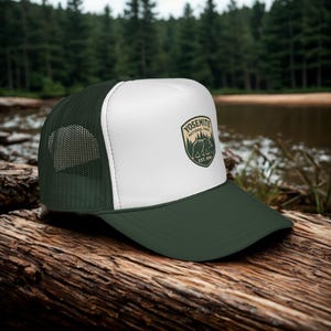 May include: A forest green and white trucker hat featuring the Yosemite National Park logo. The logo includes a bear, trees, and the text "Yosemite National Park Est. 1890". The hat has a mesh back and a curved brim.