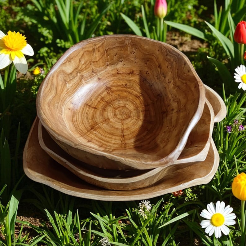 Puede incluir: Un conjunto de cuatro cuencos de madera apilados. Los cuencos muestran un patr&oacute;n de vetas de madera natural y un tono marr&oacute;n claro. Tienen una forma org&aacute;nica y r&uacute;stica. Sobre un fondo de hierba verde y flores coloridas.