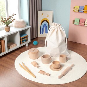 May include: A wooden toy kitchen set with a natural finish, including a pot with a lid, a cup, a bowl with a saucer, a rolling pin, and other accessories. The set is displayed on a white circular rug with a drawstring bag.