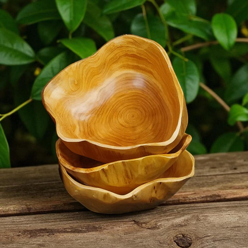 Puede incluir: Una pila de cuatro cuencos de madera con un acabado natural de color marr&oacute;n claro. Los cuencos tienen una forma org&aacute;nica e irregular y patrones de vetas de madera visibles. Est&aacute;n apilados sobre una superficie de madera con un fondo de hojas verdes.