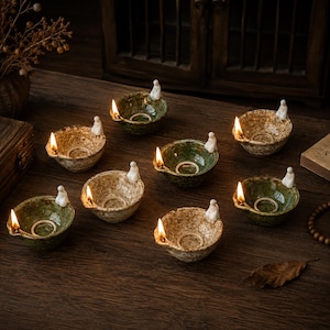 May include: A collection of eight small ceramic oil lamps with lit flames. The lamps are in shades of green and brown, each with a small white figurine attached. The lamps are arranged on a dark wooden surface.