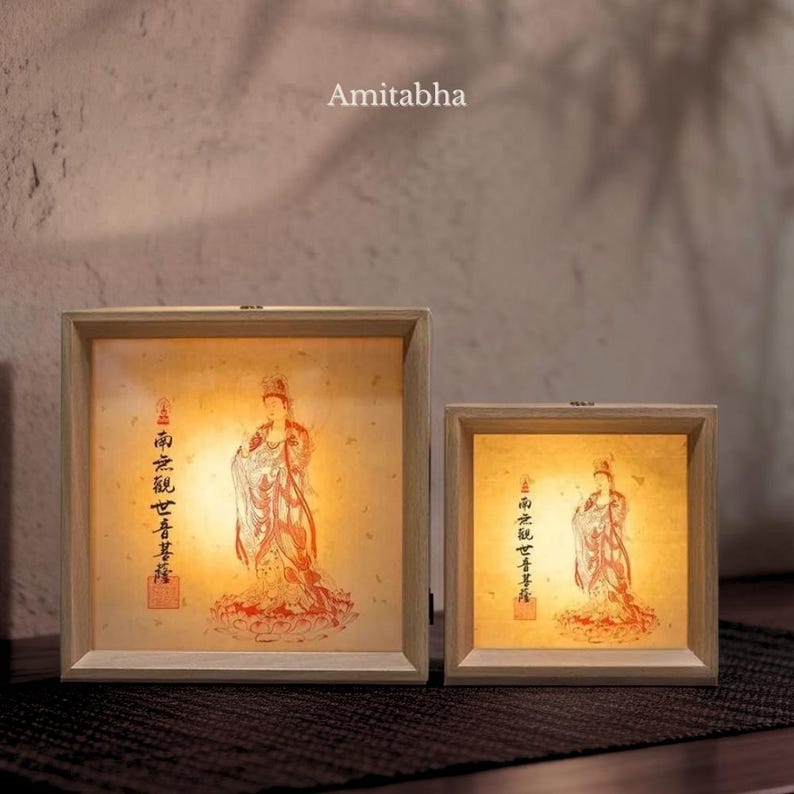 Puede incluir: Dos cajas de luz cuadradas con marcos de madera clara, cada una con una imagen c&aacute;lida e iluminada de una figura con t&uacute;nicas tradicionales, caligraf&iacute;a china y un sello rojo. La palabra Amitabha est&aacute; impresa sobre la caja de luz m&aacute;s grande.