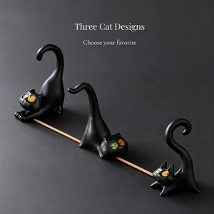 May include: Three black cat-shaped incense holders with arched tails and expressive eyes in yellow and green. The text Three Cat Designs and Choose your favorite appears above the figures, which are arranged in a line with a wooden stick.