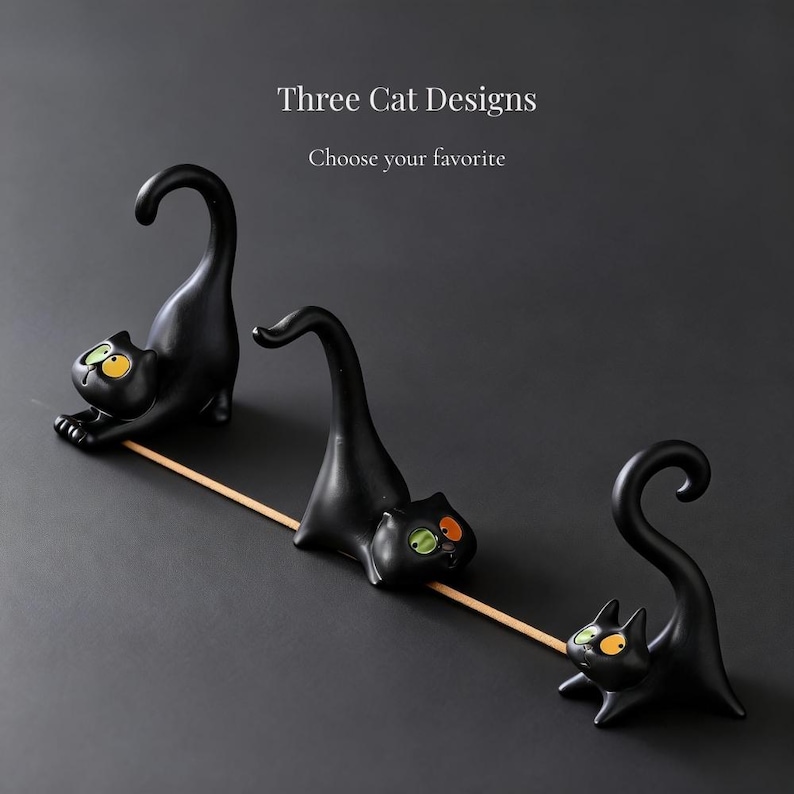 May include: Three black cat-shaped incense holders with arched tails and expressive eyes in yellow and green. The text Three Cat Designs and Choose your favorite appears above the figures, which are arranged in a line with a wooden stick.
