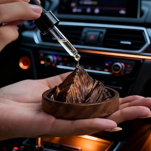 K&ouml;nnte beinhalten: Ein h&ouml;lzerner Auto-Lufterfrischer in Form einer Bergkette, gehalten in einer Hand. Eine Pipette gibt Fl&uuml;ssigkeit auf den Berg ab. Der Lufterfrischer steht auf einer runden Holzbasis. Der Hintergrund zeigt das Armaturenbrett eines Autos.