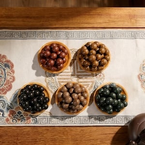 May include: Five small circular wooden bowls, each filled with a collection of smooth, spherical beads in distinct colors including dark brown, light brown, black, tan, and dark green, arranged in a cluster on a patterned fabric runner.
