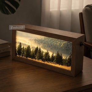 Può includere: Una scatola luminosa rettangolare in legno con un'immagine retroilluminata di un paesaggio forestale nebbioso con alberi sempreverdi e luce dorata. La cornice è in legno marrone con venature visibili. Il testo Zenboodi è in alto a sinistra.