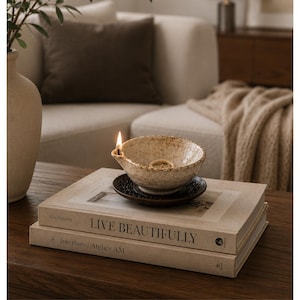 May include: A burning candle in a small, textured ceramic bowl on a dark saucer. The bowl is placed on a stack of books with the title "LIVE BEAUTIFULLY". The scene is set on a wooden table, with a neutral-toned sofa in the background.