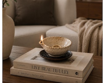 May include: A lit ceramic oil lamp with a speckled cream finish and a spout, resting on a dark brown decorative saucer. The lamp is placed on top of two stacked beige books with the text "LIVE BEAUTIFULLY" and "Interiors | Atelier AM" printed.