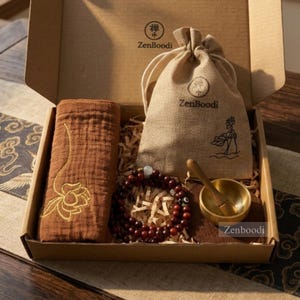 May include: A gift box containing a brown towel with embroidered flower, a drawstring bag, a wooden bracelet, a small brass bowl, and a wooden mallet. The box and bag are branded with the text "ZenBoodi".