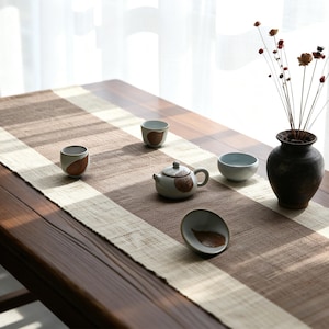 May include: A tea set consisting of a small ceramic teapot and three matching cups, each featuring a brown leaf motif. The set is arranged on a textured brown and cream table runner alongside a dark ceramic vase containing dried flowers.