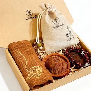 May include: A gift box with ZenBoodi branding, containing a brown towel with gold lotus embroidery, a small drawstring bag, a wooden bowl, and a beaded bracelet. The box is filled with shredded paper.