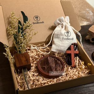 Puede incluir: Una caja de regalo con la marca ZenBoodi contiene un jarrón de madera con flores secas, una pequeña bolsa con cordón, un colgante de cruz de madera y una talla de madera con forma de montaña. La caja está llena de papel triturado.