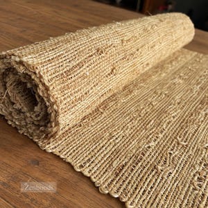 May include: A roll of natural tan woven jute fabric featuring a coarse, textured grid pattern. The material is partially unrolled, displaying a consistent, fibrous weave throughout the length of the textile. The brand name Zenboodi is visible.