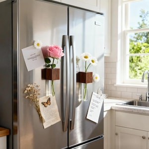 Könnte beinhalten: Edelstahlkühlschrank mit magnetischen Holzvasen mit frischen Blumen. Eine rosa Rose und weiße Gänseblümchen sind ausgestellt. Zusätzliche Magnete sind ein Schmetterling und Notizen.