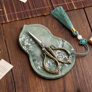 Könnte beinhalten: Dekorative Messingschere mit verzierten Griffen auf einem grünen Etui mit floralem Muster. Ein passender grüner Quastenanhänger mit dekorativer Perle und Knoten liegt neben der Schere. Die Schere ist für Bastelarbeiten oder Nähen gedacht.