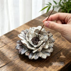 May include: A ceramic incense holder shaped like a blooming flower with marbled white, gray, and brown petals. The center of the flower features a dark, textured stamen designed to hold a thin brown incense stick upright in the center.