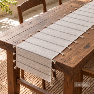 May include: A rectangular table runner featuring a cream-colored woven fabric with thin, evenly spaced horizontal dark gray stripes. The edges of the runner are decorated with small, matching cream-colored tassels along both sides.