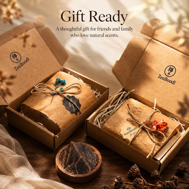 May include: Two open brown cardboard gift boxes labeled ZenBoodi, each containing a wrapped item tied with twine and dried flowers. One package has blue flowers, the other red. A small wooden bowl with dark resinous wood sits in the foreground.