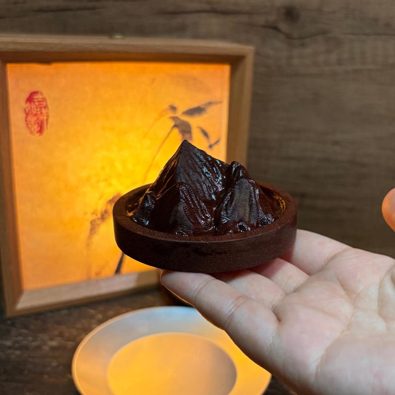 May include: A dark brown, carved wooden mountain sculpture set within a circular wooden base. The mountain features textured, jagged peaks and deep crevices, creating a realistic topographical appearance. The object is held in a palm.