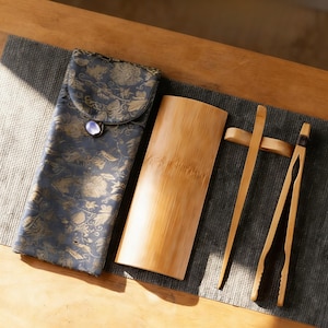 May include: An assortment of tea ceremony tools, including a patterned case, a bamboo container, and bamboo tongs. The case is dark blue with a gold floral design and a button closure. The tongs are light brown.