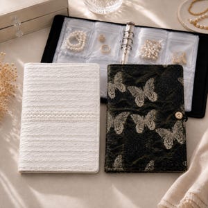 May include: Two rectangular jewelry organizers with ring-bound inserts. One organizer is white with a textured lace-like pattern, and the other is black with a silver butterfly print and a single gold-tone snap button closure.