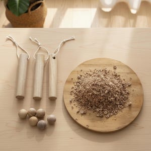 May include: A collection of wooden craft supplies on a light wood surface. The items include three cylindrical wooden handles with string, a pile of wood shavings on a round wooden board, and several small wooden spheres.