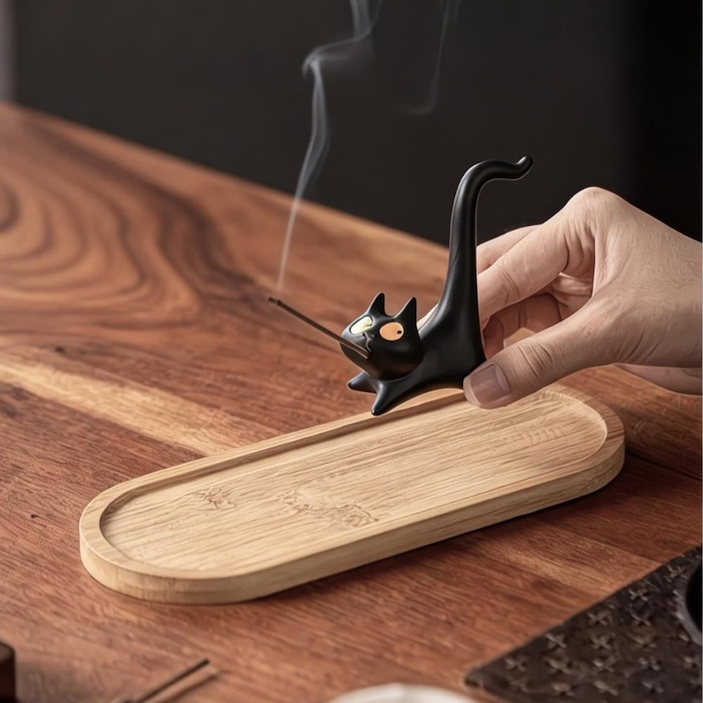 May include: A black ceramic cat-shaped incense holder with yellow eyes, featuring an upright tail and a small opening in the mouth to hold a thin incense stick. The holder is paired with an oval-shaped light brown wooden tray for ash collection.