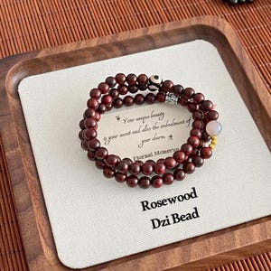 Op de afbeelding: Een rozenhouten Dzi-kralenarmband met drie strengen donkerrode kralen. De armband bevat een witte en zwarte kraal, een lichtroze kraal en een klein kaartje met de tekst "Your unique beauty is your asset and also the embodiment of your charm."