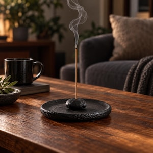 May include: A black, circular, textured incense holder with a central stone-like base holding a single, thin, light brown incense stick. A thin trail of white smoke rises vertically from the tip of the incense stick into the air.